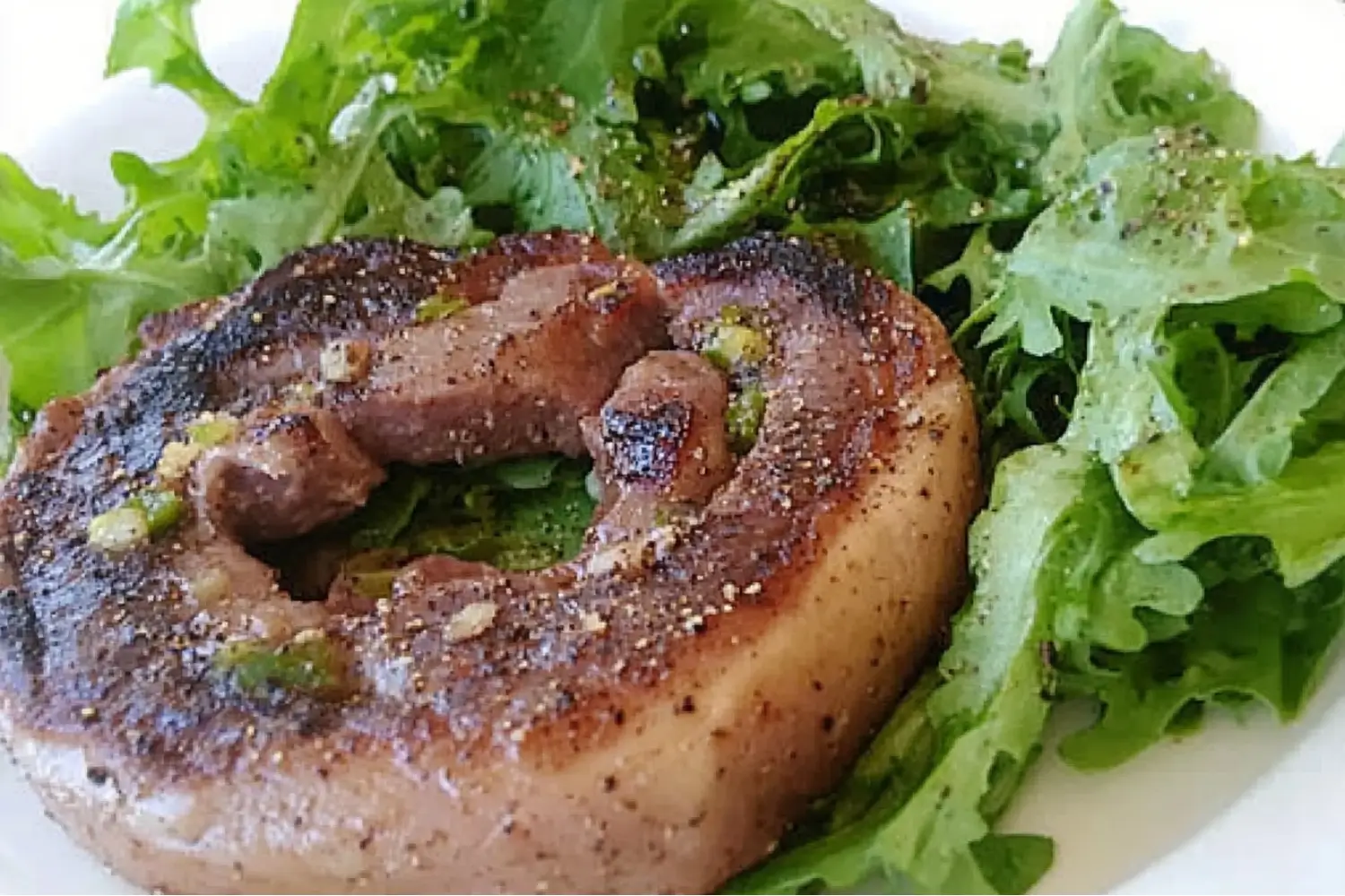 A grilled beef roll seasoned with spices and herbs, served on a white plate alongside a bed of fresh green leafy salad.