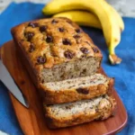 Moist protein banana bread loaf partially sliced on a wooden board with bananas and yogurt