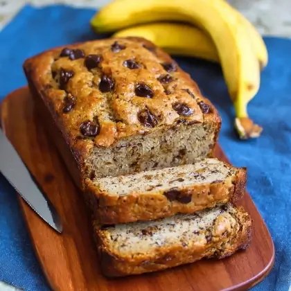 Moist protein banana bread loaf partially sliced on a wooden board with bananas and yogurt